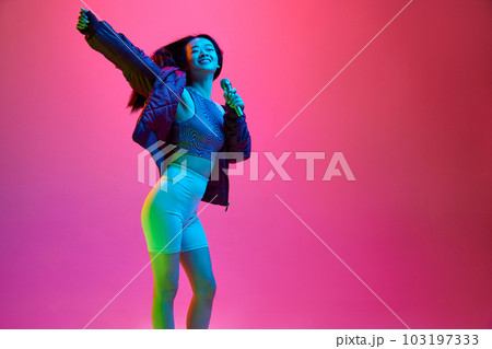 Portrait of happy, smiling, young girl singing in microphone against pink studio background in neon light. Leisure Portrait of happy, smiling, young girl singing in microphone against pink studio background in neon light. Leisure 103197333