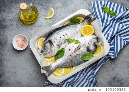 Raw fish dorado in casserole dish with ingredients lemon, fresh basil, bottle of olive oil on wooden cutting board with knife on rustic stone background top view, cooking healthy fish dorado Raw fish dorado in casserole dish with ingredients lemon, fresh basil, bottle of olive oil on wooden cutting board with knife on rustic stone background top view, cooking healthy fish dorado 103199388