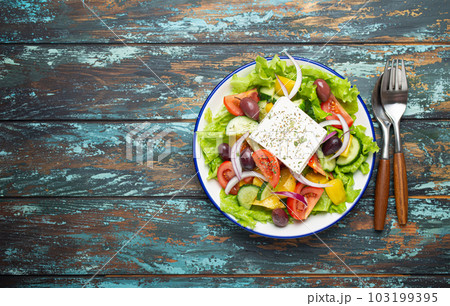 Traditional Greek Salad with Feta Cheese, Tomatoes, Bell Pepper, Cucumbers, Olives, Herbs in white ceramic bowl on blue rustic wooden table background from above, Cuisine of Greece. Copy Space Traditional Greek Salad with Feta Cheese, Tomatoes, Bell Pepper, Cucumbers, Olives, Herbs in white ceramic bowl on blue rustic wooden table background from above, Cuisine of Greece. Copy Space 103199395