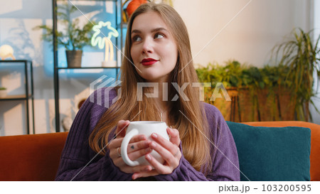Pretty smiling young woman drinking a cup of coffee or herbal tea sitting at home couch, break time Pretty smiling young woman drinking a cup of coffee or herbal tea sitting at home couch, break time 103200595
