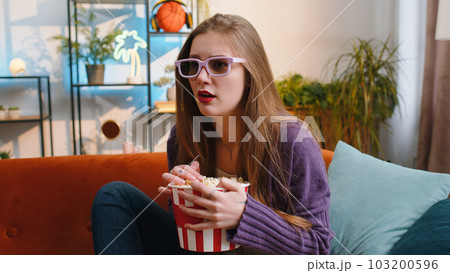 Pretty woman sits on sofa eating popcorn, watching interesting TV serial, sport game online at home Pretty woman sits on sofa eating popcorn, watching interesting TV serial, sport game online at home 103200596