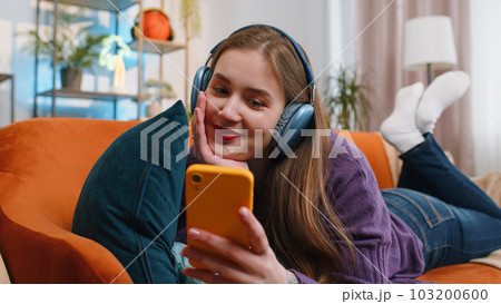 Happy young woman in wireless headphones relaxing sitting on sofa at home listening favorite music Happy young woman in wireless headphones relaxing sitting on sofa at home listening favorite music 103200600