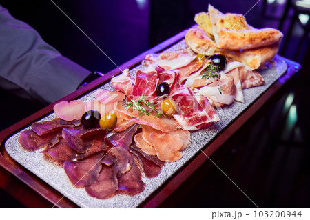 The waiter holds a wooden plate with various sliced dried meats delicacies 103200944