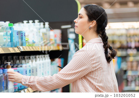 Side view of portrait of Caucasian young woman takes shampoo bottle from shelf. Choice of care cosmetics in supermarket. Concept of shopping and purchase Side view of portrait of Caucasian young woman takes shampoo bottle from shelf. Choice of care cosmetics in supermarket. Concept of shopping and purchase 103201052