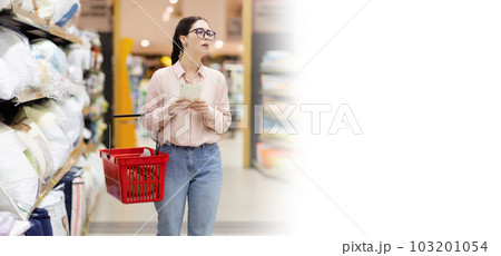 Web banner of shopping with mock up. Young caucasian woman wearing glasses holds cart and thoughtfully choises pillow and bedding. Concept of supermarket purchase 103201054
