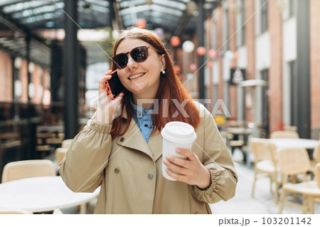 Happy cheerful young redhead woman in fashion office clothes and sunglasess talking on Phone with eco cup of tea. Mockup, Woman holding smartphone and bio coffee cup. Urban concept 103201142
