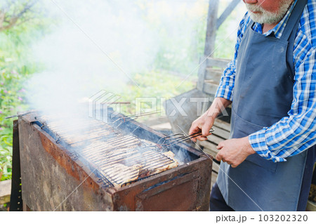 an elderly man with a beard in work clothes cooks meat steaks on the grill  103202320