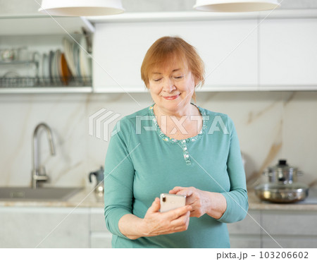 Positive mature woman in kitchen with phone in hand communicates in social network with pensioners Positive mature woman in kitchen with phone in hand communicates in social network with pensioners 103206602