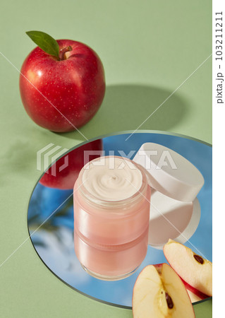 Blank label pink cosmetic container filled with cream texture placed on round mirror. Apple (Malus domestica) enhances skin condition 103211211