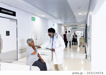 African american male doctor with tablet talking to diverse senior female patient in busy corridor 103214474