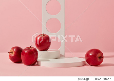 Scene for a product advertisement with a simple concept: White podiums and geometric object decorated on a pink background with some red apples. Blank space to place your product. Front view. Scene for a product advertisement with a simple concept: White podiums and geometric object decorated on a pink background with some red apples. Blank space to place your product. Front view. 103214627