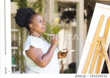 Focused happy senior african american woman holding paint palette painting canvas on easel at home Focused happy senior african american woman holding paint palette painting canvas on easel at home 103216416