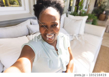 Portrait of happy senior african american woman smiling during video call in sunny living room 103216419