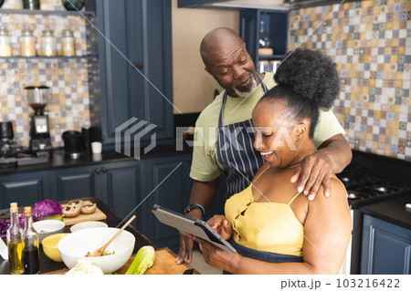 Happy senior african american couple preparing meal together, using tablet in kitchen Happy senior african american couple preparing meal together, using tablet in kitchen 103216422