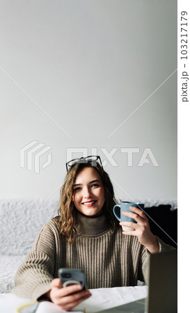 Young student taking a break from online studies, enjoying coffee and phone, laughing at memes and 103217179