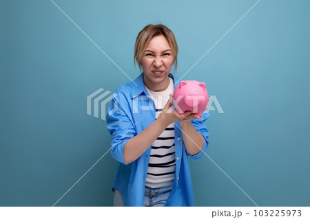 blond neat cheerful girl with a grimace holds a piggy bank on a blue background 103225973