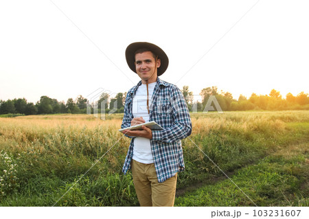 Farmer man. In the midst of a lush green wheat field, a farmer stands holding a tablet in his hands, embodying the concept of a smart farm. Sunset Farmer man. In the midst of a lush green wheat field, a farmer stands holding a tablet in his hands, embodying the concept of a smart farm. Sunset 103231607