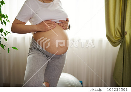 Close-up pregnant woman with naked belly, practicing yoga tree pose at home. Expectant female showing her tummy with stretch marks. Skin care. Body positivity. Pregnancy. Childbirth. Healthy lifestyle 103232591