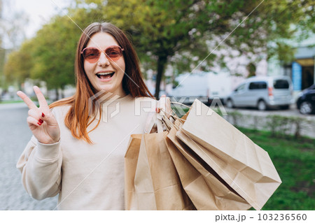 Happy young redhead woman holding shopping paper bags in hands, walking at city street, black friday. Urban lifestyle concept. Shopping banner, for mockup Happy young redhead woman holding shopping paper bags in hands, walking at city street, black friday. Urban lifestyle concept. Shopping banner, for mockup 103236560