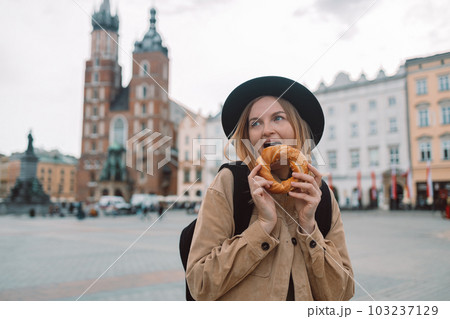 Attractive young female tourist with stylish clothes and a boho hat eating bagel obwarzanek traditional polish cuisine snack waling on Market square in Krakow. Traveling Europe  103237129
