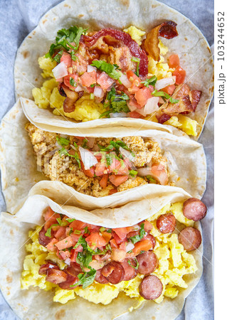 Mexican breakfast three tacos sausage bacon egg tomato chives flour tortillas food on white paper Mexican breakfast three tacos sausage bacon egg tomato chives flour tortillas food on white paper 103244652
