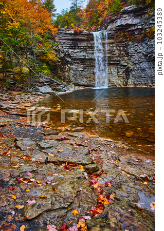 Large waterfall over cliff edge into body of water with rocks and fall leaves 103245389