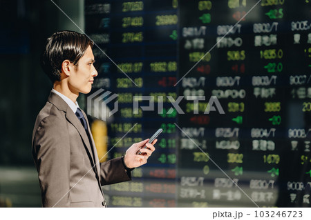 スマートフォンを手に持ち屋外の電光掲示板に映る株価ボードを自信の表情で見る30代のスーツ姿の男性 103246723