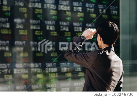 ビジネス街に設置された屋外電子掲示板の株価ボードを見る30代の日本人のビジネスマンの後姿 103246730