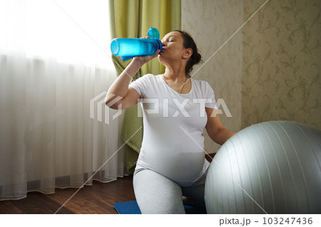 Multi-ethnic pregnant woman practices yoga and prenatal stretching exercises with a fit ball at home, drinks water from bottle, sitting on a blue mat, renewing aqua balance after pregnancy fitness 103247436