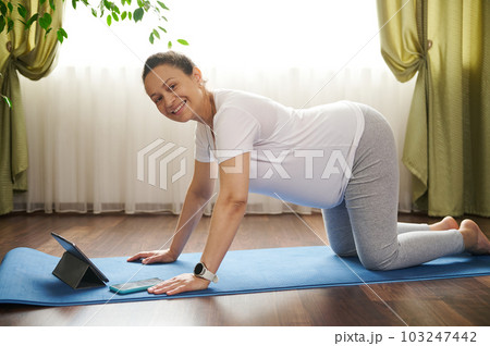 Happy pregnant woman in white t-shirt and grey leggings, standing at table pose on hands and knees on fitness mat, practicing prenatal gymnastics and stretching exercises, using digital tablet at home 103247442