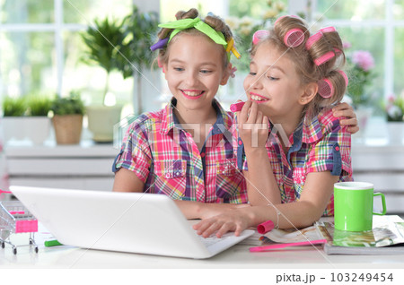 Cute twin girls in hair curlers with laptop at home appying lipstick 103249454