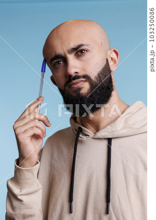 Puzzled arab man in thoughts holding pen to head while looking away. Young bald bearded person rubbing temple while planning and solving problem with uncertain expression Puzzled arab man in thoughts holding pen to head while looking away. Young bald bearded person rubbing temple while planning and solving problem with uncertain expression 103250086