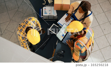 African american people looking at supplies list on pc, working with computer to check inventory and logistics. Employees organizing merchandise on shelves in warehouse space. Handheld shot. 103250218