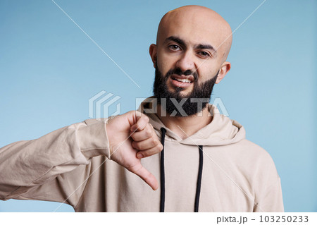 Arab man using thumb down gesture to show disapprovement while looking at camera with negative emotion grimace. Young adult person expressing disagreement studio portrait Arab man using thumb down gesture to show disapprovement while looking at camera with negative emotion grimace. Young adult person expressing disagreement studio portrait 103250233
