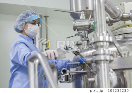 Pharmaceutical Female Worker Wearing Protective Clothing in Sterile Environment and Standing Next to Manufacturing Machine at Pharmaceutical Plant Pharmaceutical Female Worker Wearing Protective Clothing in Sterile Environment and Standing Next to Manufacturing Machine at Pharmaceutical Plant 103253239