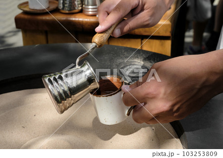 Barista pours freshly made Turkish coffee on the sand in a cezve into a cup, close-up. A professional barista prepares coffee by hand in the traditional way. Selective focus, space for text Barista pours freshly made Turkish coffee on the sand in a cezve into a cup, close-up. A professional barista prepares coffee by hand in the traditional way. Selective focus, space for text 103253809