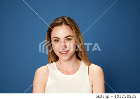 Portrait of young, beautiful, blonde girl posing, looking at camera with smile against blue studio background. Happiness Portrait of young, beautiful, blonde girl posing, looking at camera with smile against blue studio background. Happiness 103258228