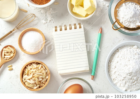 Notepad and pen for recipe with baking or cake ingredients in bowls, butter, milk, nuts and eggs with gold whisk on marble table 103258950
