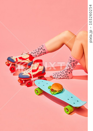 Female legs in vintage rollers and funny socks with delicious burger on skateboard against pink background. Delivery 103260254