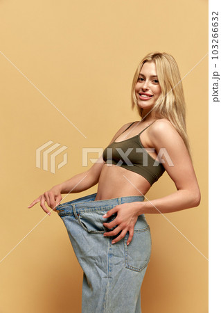 Young beautiful woman with slim body, posing in giant, oversized jeans against yellow studio background. Weight loss, sport and diet Young beautiful woman with slim body, posing in giant, oversized jeans against yellow studio background. Weight loss, sport and diet 103266632
