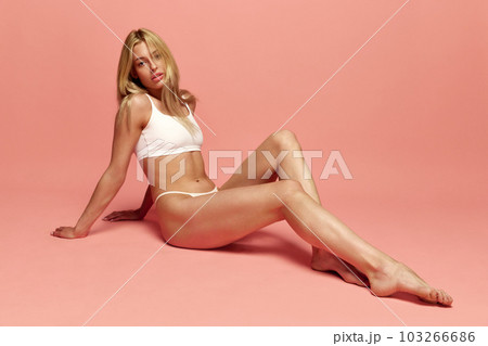 Portrait of young beautiful, blonde girl posing in white underwear against pink studio background. White, slim body Portrait of young beautiful, blonde girl posing in white underwear against pink studio background. White, slim body 103266686