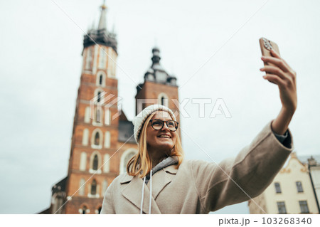 Young smile woman tourist In a stylish coat takes selfie on mobile phone on the market square in Krakow in Poland Young smile woman tourist In a stylish coat takes selfie on mobile phone on the market square in Krakow in Poland 103268340