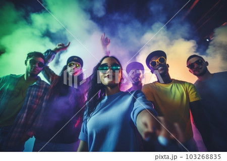 Group of young people wearing neon clothes and sunglasses dancing at a rave party in front of a DJ booth with colorful lights and smoke effects in the background. Group of young people wearing neon clothes and sunglasses dancing at a rave party in front of a DJ booth with colorful lights and smoke effects in the background. 103268835
