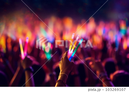 A close-up shot of hands holding glow sticks in the air at a rave party with a blurry crowd of people dancing in the background. Generative Ai 103269107