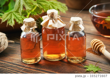 Three bottles of maple syrup or healthy tincture and spring maple leaves. Bowl of maple syrup and wooden dipper on background. 103271607