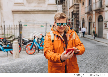 Cheerful young guy walking on city street with modern smartphone in hands. Handsome man in sunglasses reading some good and smiling. Technology for lifestyles. 103287587