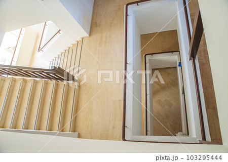 Hotel staircase with slightly curved architecture, windows and historic metal railing. Stairs are tiled and the handrail is supplemented with wooden handrail. There is also wood on walls and paneling. 103295444