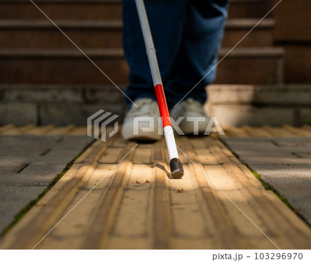 A blind woman walks outdoors using a cane along a tactile yellow tile. 103296970