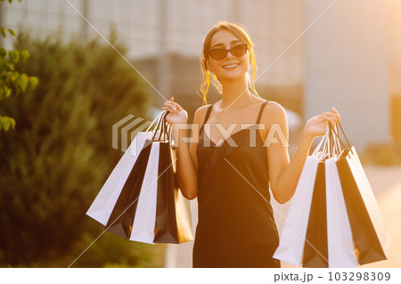 Elegant brunette woman wears black dress holding black and white shopping bags at sunset. Black friday, sale. 103298309