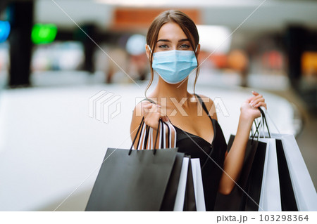 Beautiful fashionable woman in protective medical mask with shopping bags walking near mall. Covid-19. 103298364